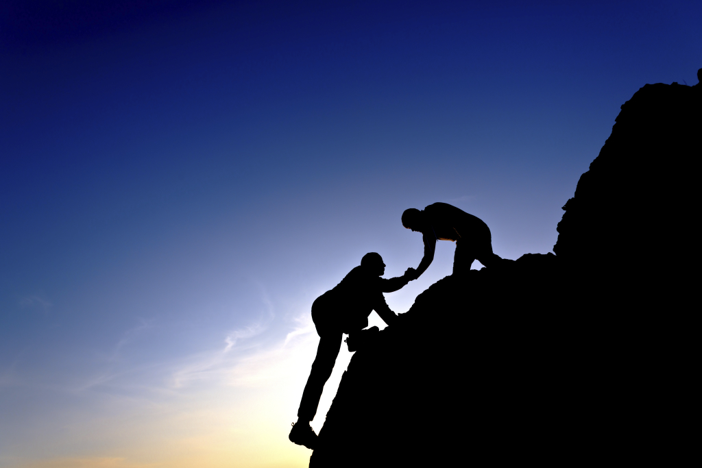 One person helping another person climb up a steep rocky crag.